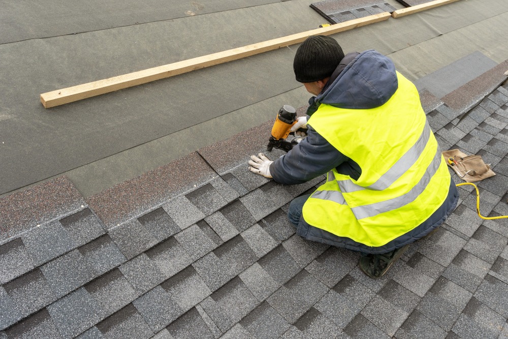 Qualified workman in uniform work wear using air or pneumatic nail gun and installing shingles