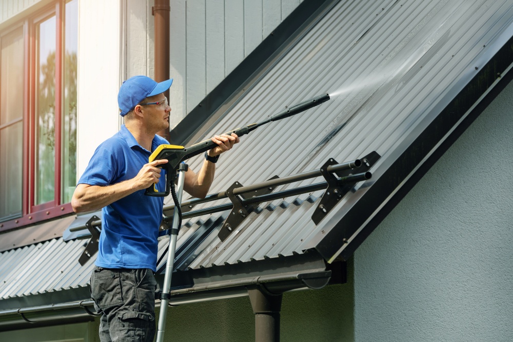 Person standing on ladder and cleaning house metal roof with high pressure washer